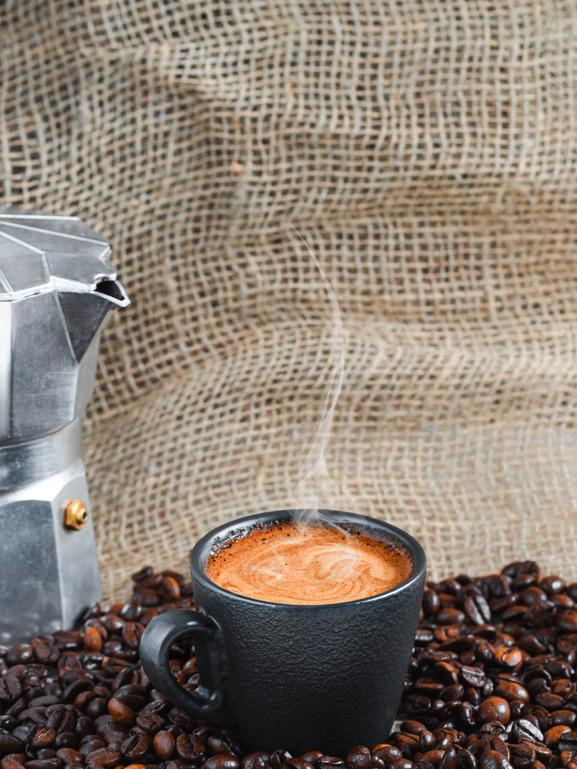 Cup of strong aromatic espresso coffee with froth among roasted coffee beans, next to a coffee pot. Steam rises above a cup of coffee. Close-up, copy space with burlap on the background